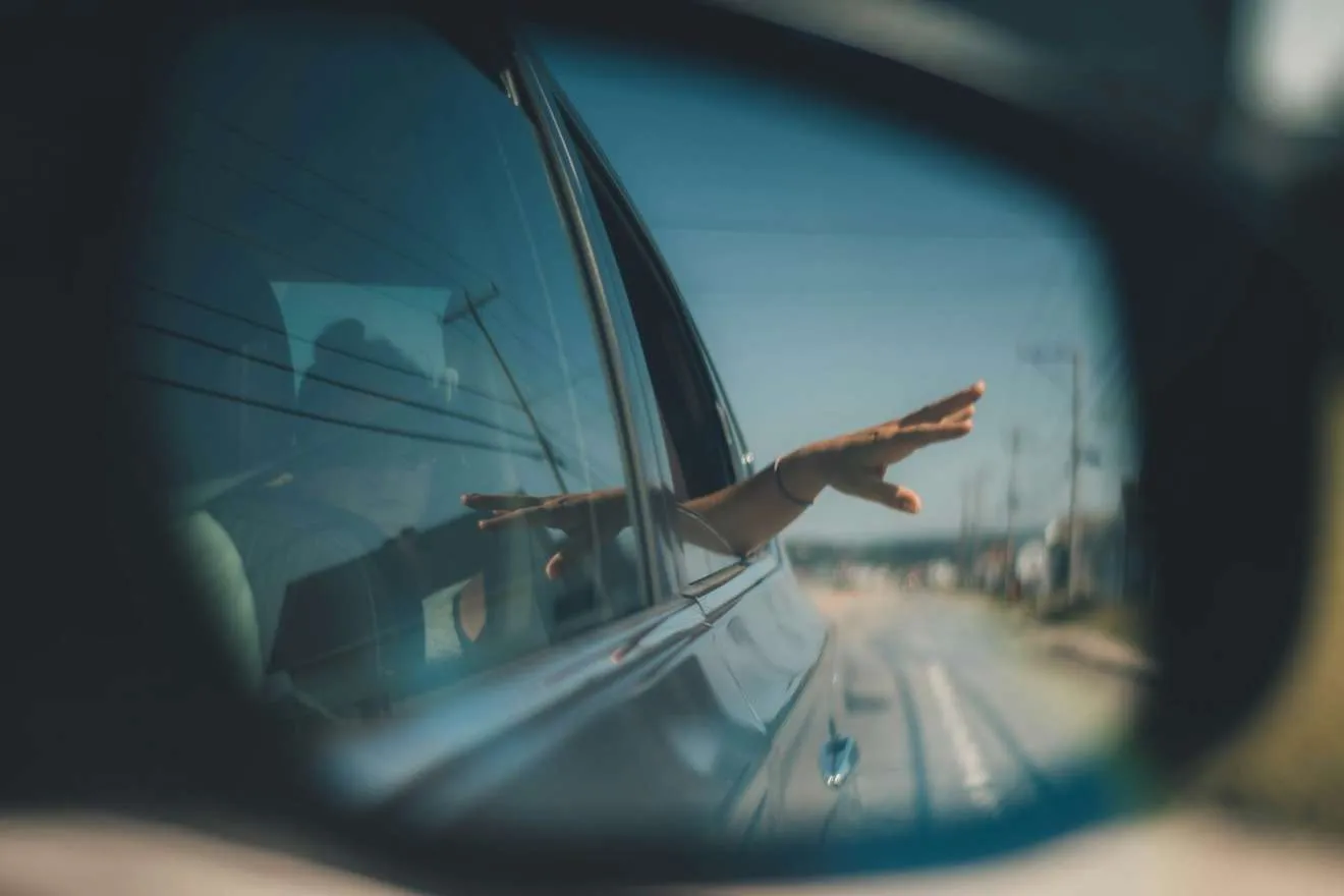 A passenger's hand out of a car window, seen in the wing mirror on a sunny road